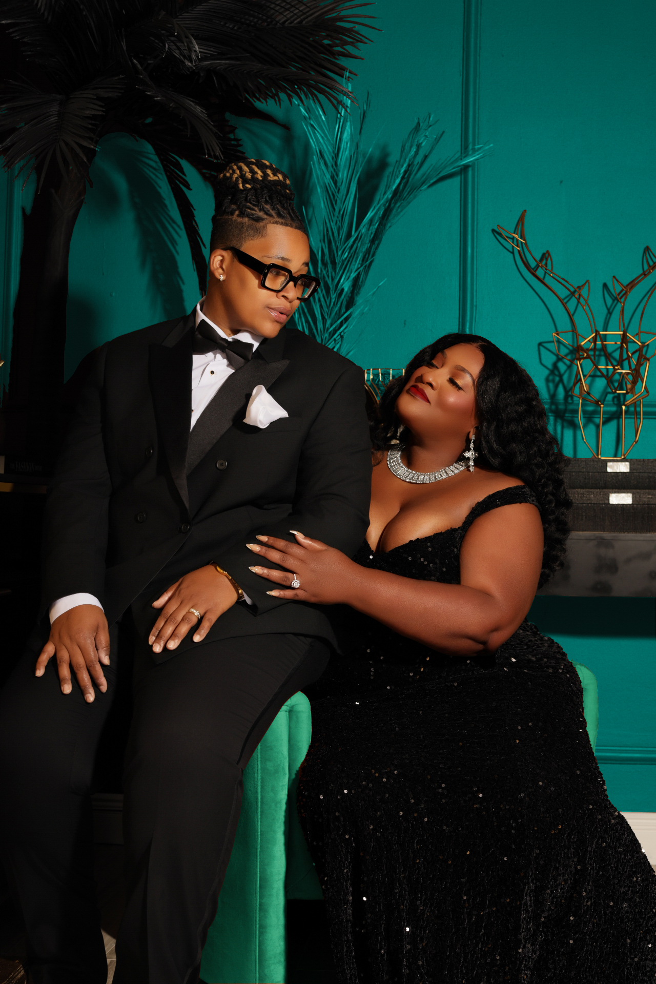 The couple seated together in black formalwear against a teal backdrop
